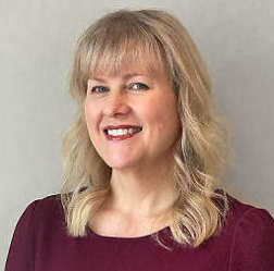 A woman with blonde hair wearing a maroon top, smiling and looking towards the camera