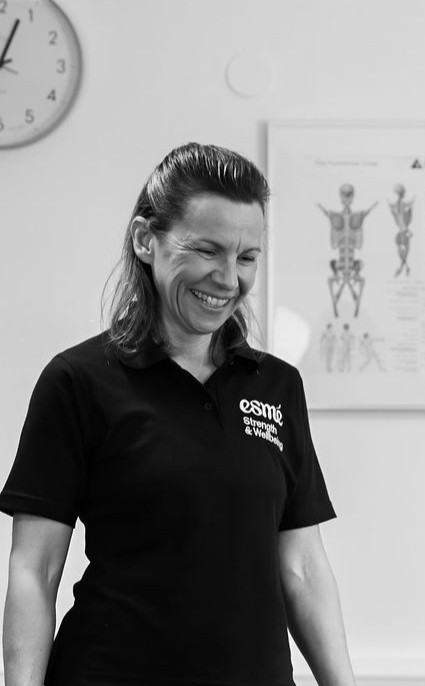 A woman in a black polo with the esm? Strength & Wellbeing logo smiles while looking down, standing in a clinic space with an anatomical poster and a wall clock behind her.