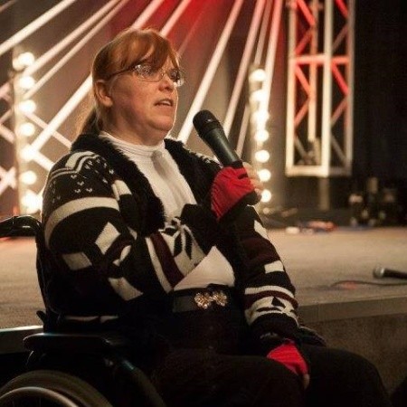 The image captures Fiona (Fee) Stevenson sitting in a wheelchair on a stage, holding a microphone. Fee is dressed in a black and white patterned sweater, a white shirt, and red gloves. The stage is brightly lit, with metal trusses framing the background, suggesting that this might be a performance or public speaking event. There's a sense of engagement and presence, as the Fee appears to be actively participating in the moment.