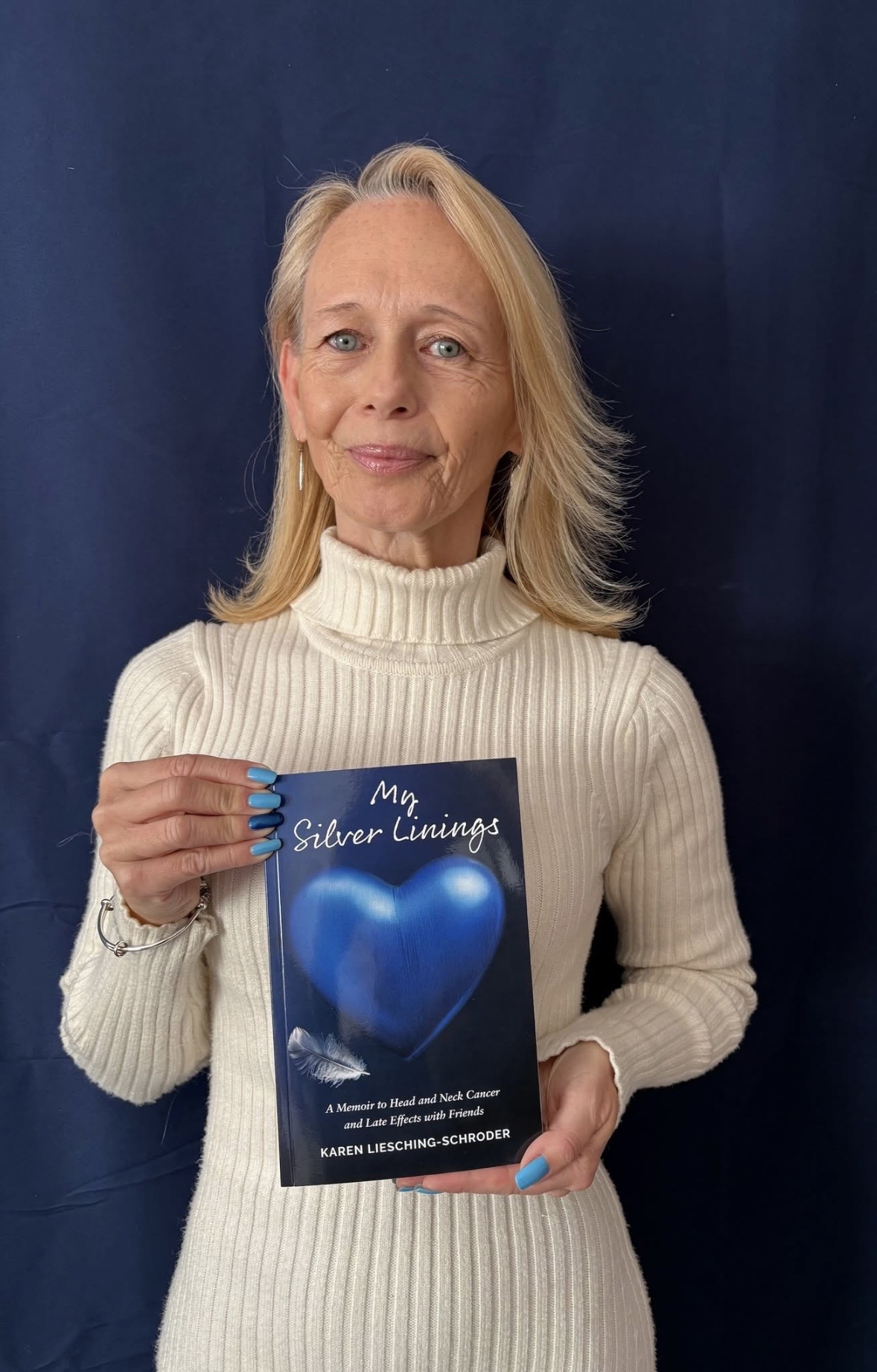 Karen Liesching-Schroder stands against a dark blue background, wearing a cream-coloured ribbed turtleneck sweater. Karen holds her book titled My Silver Linings. The cover features a large blue heart with a white feather resting across it. "A Memoir to Head and Neck Cancer and Late Effects with Friends" appears in the subtitle. Karen Liesching-Schroder's name appears at the bottom of the cover. The overall mood of the image is calm and reflective, highlighting a memoir about resilience and life after cancer.
