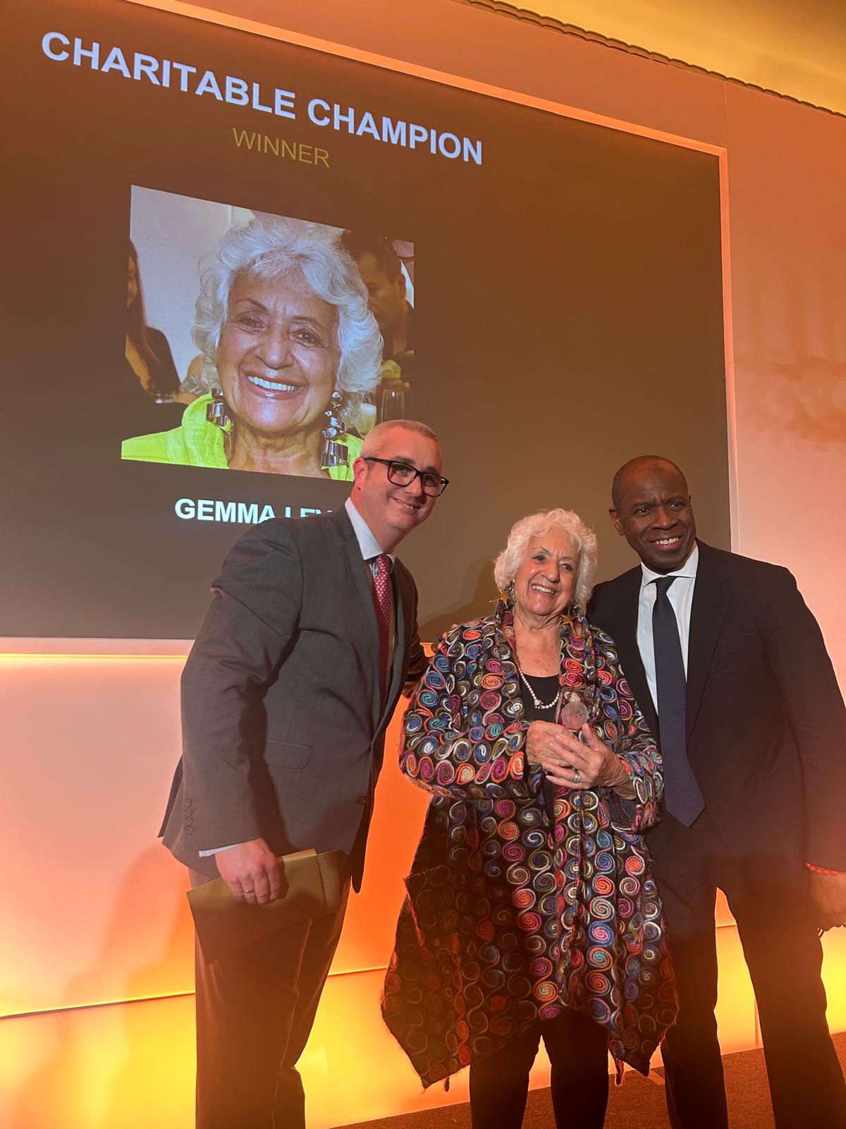 "Gemma Levine stands between two men at an award ceremony, smiling as she receives the 'Charitable Champion' award. All are dressed formally. Behind them, a large screen displays her photo, the title 'Charitable Champion,' the word 'Winner,' and her name. The setting is warmly lit in orange tones, creating a celebratory atmosphere."